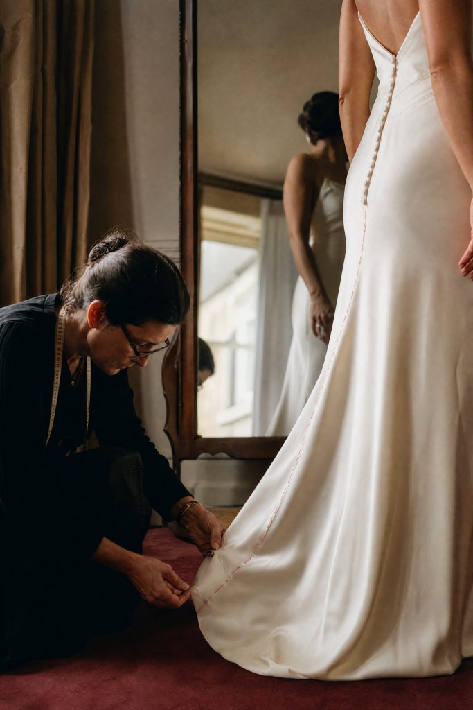 Une couturière agenouillée épingle l’ourlet d’une robe en satin ivoire, devant un miroir